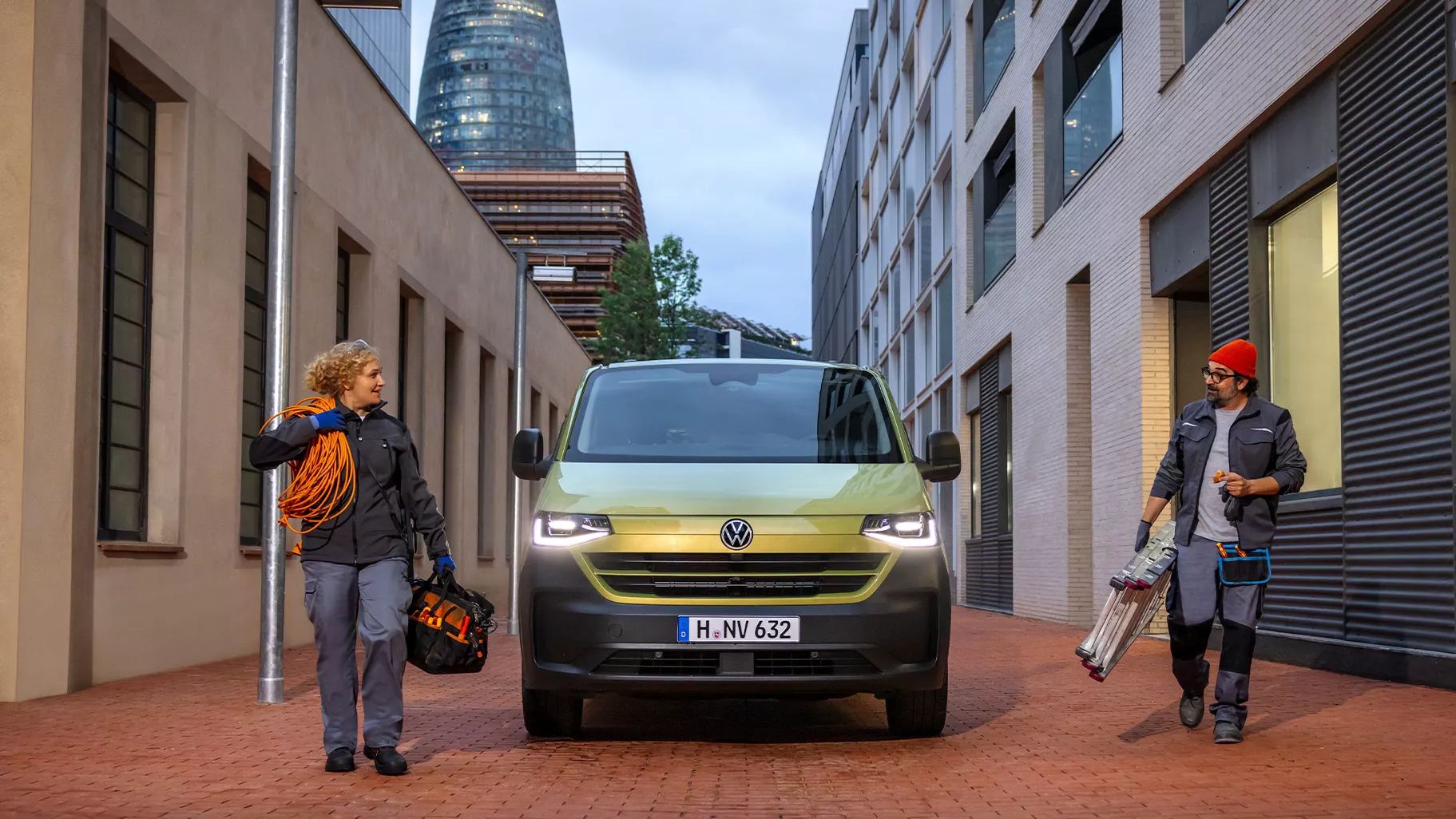 Der neue Transporter in grün, steht zwischen modernen Gebäuden auf gepflasterter Straße. Zwei Handwerker stehen links und rechts vom Auto; einer trägt Kabel, der andere eine Leiter. Im Hintergrund ist ein markantes, eiförmiges Gebäude zu sehen.