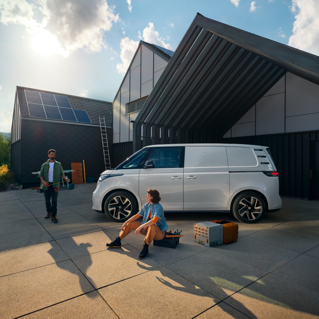 Ein weißer Volkswagen ID. Buzz Cargo vor einer Werkshalle, im Vordergrund sind ein Mann und eine Frau zu sehen