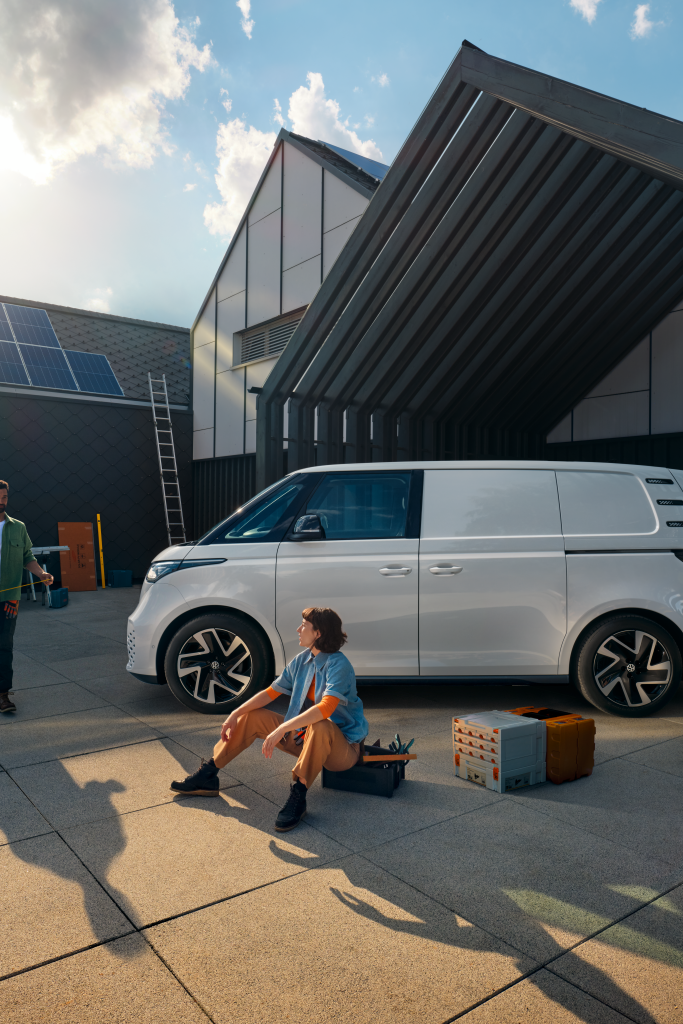 Ein weißer Volkswagen ID. Buzz Cargo vor einer Werkshalle, im Vordergrund sind ein Mann und eine Frau zu sehen
