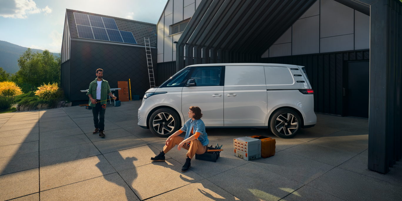 Ein weißer Volkswagen ID. Buzz Cargo vor einer Werkshalle, im Vordergrund sind ein Mann und eine Frau zu sehen